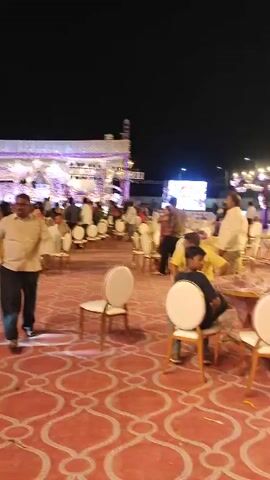The video depicts an outdoor evening event, likely a wedding reception or large celebration, in Askihala, India. The venue is elaborately decorated under the night sky with a festive atmosphere.

The ground is covered with a vibrant red carpet featuring a repeating white geometric pattern. Numerous white chairs with golden frames are arranged throughout the space, some placed around tables where guests are seated, and others freestanding. In the background, a large stage or tent structure is visible, adorned with white fabric drapes, string lights, and spotlights that brightly illuminate the area. Other decorative elements include archways and light fixtures.

Various people are seen moving and interacting within the venue. At the beginning, a man in a beige shirt and dark pants walks across the patterned carpet towards the camera, while other guests are visible mingling in the background. The camera then pans to reveal more of the crowded event space, with people sitting at tables and additional decorative lighting. Towards the end, a woman wearing a flowing blue and white patterned dress quickly runs from left to right across the foreground. The overall scene suggests a lively and well-attended social gathering. There is no visible text in the video.