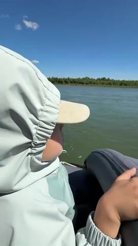 A first-person perspective video shows a person on a boat sailing on a river. The person is wearing a light blue hooded jacket and a beige cap. The water is green and calm, and the sky is blue with a few white clouds. The background shows a lush green forest on the riverbank. The camera pans slightly to show the water and the surrounding landscape.