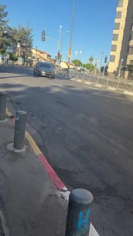 The video shows a black pickup truck driving on a city street. The road surface appears worn and patched with black marks.  On the left side of the road, there is a sidewalk with grey bollards. The curb is painted with a red, yellow, and white stripe. Streetlights and traffic signals are visible along the street. The sky is clear and blue. Buildings line the street. The truck passes by the camera, heading away from the viewer.