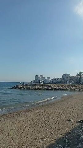 This video captures a serene beach scene in Mezitli, Turkey. The clear blue sky suggests a pleasant, sunny day, likely in the morning or late afternoon given the soft lighting. The camera pans across a sandy and pebbly beach, with gentle waves lapping at the shore. In the distance, a rocky jetty extends into the sea, with a few people visible on it, possibly fishing or enjoying the view. Further along the coastline, a cluster of modern buildings indicates a populated area or town.

The foreground of the beach shows some natural debris, like dried branches and scattered pebbles, alongside a few pieces of litter, suggesting it's a public space. The main focus of activity shifts to a group of people sitting on the beach near the water's edge. Several individuals are dressed in white, possibly for religious or cultural reasons, and they appear to be in a relaxed, contemplative state. One person is partially submerged in the water, while another is swimming further out. The overall atmosphere is one of tranquility and leisure, with people enjoying the natural beauty of the coastal environment. There is no visible text in the media file.