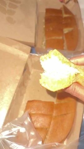 Here is a description of the video:
The video shows a hand holding up a piece of sliced terang bulan, also known as martabak manis, over two cardboard boxes containing more slices of the same dessert. The terang bulan appears to have a generous filling of cheese. The hand pulls apart the piece, revealing the cheese. The boxes sit on a blue and white patterned bedspread. The terang bulan slices are arranged in segments in the boxes. One of the boxes is partially covered with a clear plastic bag.