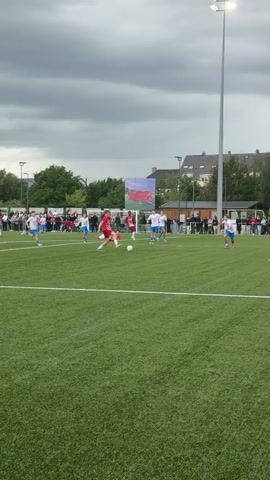 A short video captures a boy in a red and white jersey playing soccer on a green field. He dribbles the ball around another boy in a blue and white jersey and scores a goal. Spectators watch from behind a fence in the background, and buildings and trees are visible beyond them under a cloudy sky.