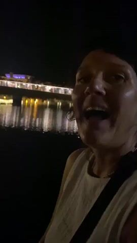 This is a short, slightly blurry video taken at night. The primary subject is a woman with dark hair, looking towards the camera and smiling. She appears to be wearing a light-colored, textured top and a dark strap across her chest. Her face is well-lit, highlighting her smile.

In the background, across what appears to be a body of water, there is a building or structure with numerous lights, reflecting on the water's surface. The overall scene is dark, indicating it is nighttime. The presence of lights and the reflections suggest an urban or populated waterfront setting.

The woman's expression and the context of the lights in the background suggest a pleasant, possibly festive, atmosphere. The activity appears to be casual, perhaps a person taking a selfie or recording a short video clip while out at night. There is no visible text in the media file. The context of Tavira, Portugal, places this scene in a historic Portuguese town often characterized by its riverfront and illuminated architecture at night.