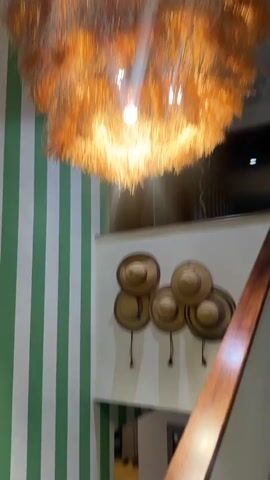 Two men walk up a beige-colored staircase with a wooden handrail. One man is wearing a black jacket, black pants, and black shoes with white socks. The other man is wearing a white and green striped jacket, striped shorts, and athletic shoes. They appear to enter a doorway at the top of the stairs. The wall is striped green and white. On the wall is a display of five straw hats. A decorative light fixture hangs from the ceiling.