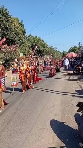 The video shows a parade or procession on a paved road under a clear blue sky. Several people are walking in the procession, dressed in elaborate, traditional costumes, predominantly red and gold. The costumes include ornate headpieces, long flowing robes, and decorative sashes. Some individuals are carrying banners or staffs. Indonesian flags are visible, mounted on poles along the side of the road and also carried by participants, indicating a possible national celebration. On the sides of the road, there are trees, some with red and white decorations, and a crowd of onlookers can be seen. The overall atmosphere appears festive.