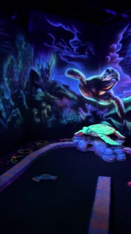 The video shows a section of glow-in-the-dark miniature golf. The walls are painted with a vibrant ocean scene in blacklight colors, with large images of sea turtles. In the foreground is the mini-golf course, with a small obstacle made of a turtle resting on rocks. The course has various geometric shapes and a faux seashell decoration. The scene is very dark, illuminated mainly by the blacklights that make the artwork glow. The background music is barely audible.