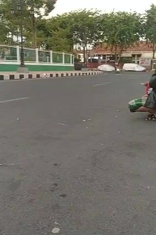 The video shows a street scene. In the foreground, a person is crouching on the asphalt. A motorcycle with its headlight on is visible in the middle distance on the road, approaching from the left. On the left side of the road, there is a sidewalk with a decorative fence and trees behind it. A lamppost is also visible. In the far distance, buildings with red roofs can be seen. The road appears to be in a somewhat neglected state with some debris scattered on it.