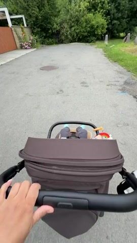 The video shows a first-person perspective of someone pushing a stroller. The stroller contains a baby wearing a plaid shirt, grey leggings, blue socks, and a light-colored baseball cap. The baby has a pacifier in its mouth and a toy attached to the stroller's handlebar. The stroller is moving along a paved path, with greenery and trees visible on either side. The sky is overcast.