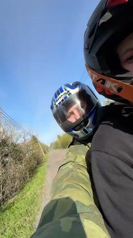 Two individuals are riding a moped or similar vehicle on a path alongside a wooded area under a clear blue sky. The camera's perspective is from behind the driver, capturing the back of their head and the face of the passenger. The driver wears a camouflage jacket, while the passenger is in a black hoodie. Both wear helmets; the driver's is blue and silver with the letters "MDS," and the passenger's is a black and orange "ATAKI" helmet with goggles. The passenger briefly flashes a peace sign to the camera. Power lines are visible above and beside the path. The vehicle is moving, as the landscape scrolls past.
