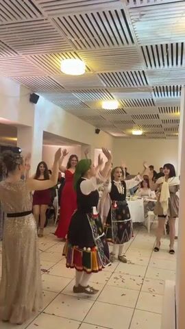 A group of women are dancing in what appears to be a banquet hall. Several of the women are wearing formal dresses in red and gold. Two of the women are wearing traditional Bulgarian clothing consisting of black vests and skirts with colorful patterns and fringed hems. They are also wearing headscarves and wooden shoes. The women all have their arms raised in the air. There is confetti on the floor. In the background, there are tables with people seated at them. The room has a dropped ceiling with fluorescent lights.