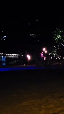The video shows a fire dancer performing on a beach at night. The dancer is silhouetted against the lights, and is surrounded by palm trees. There are people watching in the background. The water is visible in the distance, with lights reflecting on it.