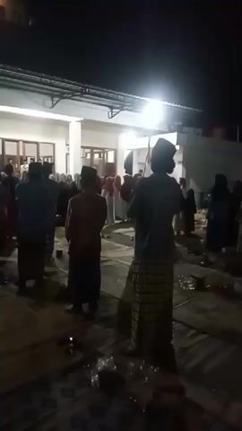 The video shows a group of people gathered outdoors at night. The scene appears to be a religious or cultural event. Many of the individuals are wearing traditional Islamic clothing, including peci (caps) and sarongs. They are standing on mats or woven rugs laid out on the ground. In the background, there is a building with an open-air covered walkway. Several lights illuminate the area, casting shadows and highlighting the figures. Some people are facing a central point, possibly listening to a speaker or participating in a prayer or ceremony. There are some plastic bottles and other discarded items visible on the ground. The overall atmosphere seems solemn and reverent.