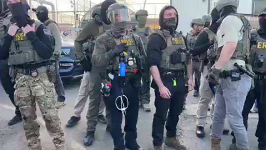 A group of approximately a dozen individuals, identified as U.S. Immigration and Customs Enforcement (ICE) agents by visible "POLICE" and "ICE" patches on their vests, are standing together outdoors. They are equipped in full tactical gear, including bulletproof vests, helmets (some with visors), and balaclavas or face masks that obscure their faces. Their attire varies, featuring camouflage pants, dark uniforms, and various tactical pouches and accessories.

They are positioned on an asphalt or concrete surface marked with a yellow line. In the background, a chain-link fence and a light-colored building or trailer are visible, suggesting an industrial or secured compound environment. The lighting indicates it is daytime, possibly late afternoon. The individuals are mostly stationary, appearing alert and in a state of readiness. One agent is seen adjusting their face covering. The scene depicts a gathering of law enforcement personnel in Newark, United States.