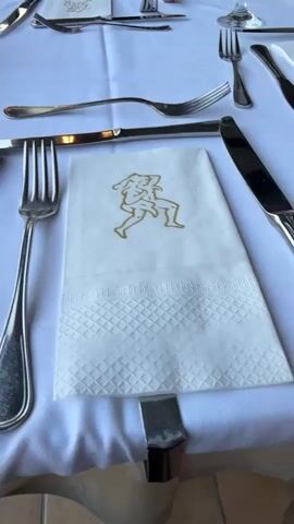 The video opens with a close-up shot of a table setting at a restaurant. There is a white tablecloth, silverware, and a folded white napkin with a gold emblem of a dancing figure. The camera then pans to the left, revealing more of the table setting, including a wine glass and a small table number that reads "66". The scene then shifts to an outdoor setting, with a view of a swimming pool and lounge chairs. The pool is a vibrant blue, and the lounge chairs are arranged on a green lawn. In the background, there are palm trees and the ocean. The lighting suggests it is daytime, possibly late afternoon, with a clear sky and bright sunshine. The overall atmosphere appears relaxed and vacation-like.

**Notable Details:**

*   **Scene:** A restaurant table setting and an outdoor poolside area.
*   **Objects:** Silverware, white tablecloth, white napkin with gold emblem, wine glass, table number "66", lounge chairs, swimming pool, palm trees.
*   **Location Cues:** The presence of a pool, lounge chairs, palm trees, and the ocean strongly suggest a resort or hotel environment in a warm climate. The table number also indicates a dining establishment.
*   **Time of Day/Weather:** Bright sunlight and clear skies suggest daytime, likely with pleasant weather.
*   **Emotions/Atmosphere:** The setting evokes feelings of relaxation, vacation, and a pleasant dining experience.

**Visible Text:**

*   Table number: "66"

**Activity/Event:** The video appears to be documenting a dining setup at a restaurant, possibly in a resort setting, and then showcasing the amenities of the location. It could be a personal video documenting a meal or a promotional clip for the establishment.