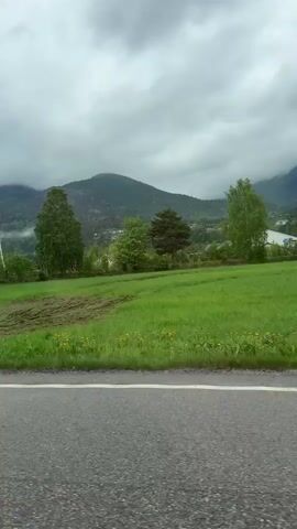 The video shows a red car driving on a road. The road has a white line marking the edge. There is a green field to the right of the road, with a mountain in the background. The sky is cloudy. A pedestrian overpass is visible.