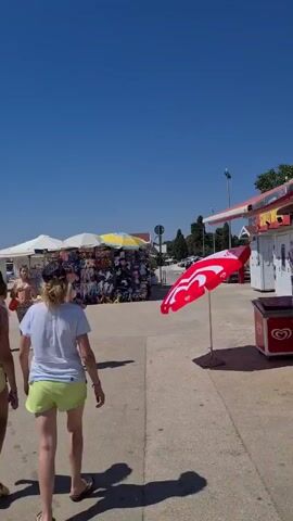 This video captures a sunny day in Općina Fažana, Croatia, likely in the late morning or early afternoon given the bright sunlight and clear blue sky. The scene appears to be a tourist area near the coast, with several market stalls set up.

In the foreground, two women are walking away from the camera, both wearing casual summer attire. The woman on the left has long, dark hair and is wearing a black baseball cap, a black top, and light yellow shorts. The woman on the right has blonde hair, is wearing a blue baseball cap, a white t-shirt, and light yellow shorts. They are walking on a paved surface, possibly a promenade or walkway.

Behind them, several market stalls are visible. Some are covered by white or yellow umbrellas, suggesting they are selling goods under the sun. The stalls are stocked with various items, including what appear to be bags, sunglasses, and other souvenirs, common in tourist destinations. One stall has a vibrant display of colorful bags. In the distance, the sea is visible, further confirming the coastal setting.

The overall atmosphere seems relaxed and typical of a summer vacation day. The weather is clearly warm and pleasant, indicated by the clear sky and the clothing worn by the people. The location context of Općina Fažana, Croatia, aligns with the visual cues of a seaside town with tourist amenities. There is no visible text in the video.