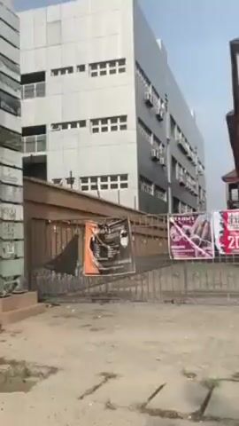 The video shows a car driving past various buildings, including what looks like an Institute of Child Health University College Hospital and other structures with fences and gates. The buildings are modern in design, mostly painted in white and gray. The video is taken from inside the moving vehicle, giving a view of the roadside architecture.