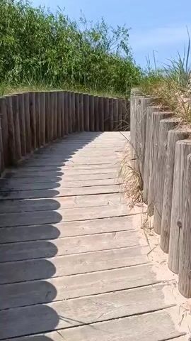 The video shows a wooden boardwalk leading through sand dunes towards a beach and the ocean. The boardwalk is lined with wooden posts. Green vegetation is visible on either side of the boardwalk. The sky is blue with some clouds. The ocean is visible in the distance, with gentle waves.