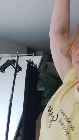 In this video, a woman with blonde, curly hair is filmed from the chest up. She is wearing a yellow t-shirt with black text that appears to be in a script font. The text reads, "The soul that makes you what you are". She is smiling and making a kissing face while raising her right arm. In the background, a clothing rack with black hangers and black clothing can be seen, as well as a large potted plant, a mirror, and a fan. The lighting suggests it is daytime, and the setting appears to be indoors, possibly a dressing room or bedroom.