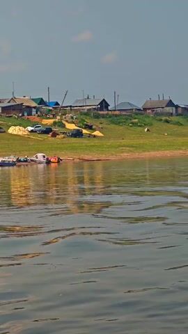 The video shows a view from a body of water, looking towards a shoreline with a small village. Several boats are docked along the shore, and a few cars are visible. The village consists of low-rise wooden buildings, with some structures appearing to be under construction or renovation. The landscape is green and hilly, with scattered trees. The sky is partly cloudy, and the sun is shining, creating reflections on the water. The overall impression is one of a peaceful rural setting.