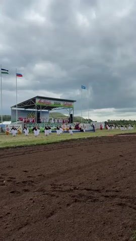 The video shows an outdoor event. In the foreground, there is a large area of dark, tilled soil. In the midground, a stage is set up with a banner that reads "Казырбас 2023". A group of people, dressed in white with black accents and holding white flags or fabric, are performing a choreographed dance in front of the stage. There are also other people on and around the stage, some appearing to be performers and others possibly organizers or spectators. Behind the stage, there are hills and trees, indicating a rural or semi-rural setting. The sky is cloudy. Three flags are visible to the left of the stage: one is green, white, and blue; another is red, white, and blue (the Russian flag); and a third is blue with a white symbol. Another blue flag with a white symbol is visible to the right of the stage. The overall atmosphere appears to be festive or ceremonial.