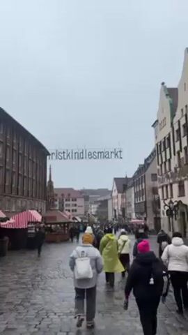 The video shows a view of a crowded street in Nuremberg, Germany. The street is lined with buildings on either side. Above the street hangs a sign that reads "Christkindlesmarkt." People are walking along the cobblestone street, past market stalls with red and white striped canopies. The sky is overcast and gray.