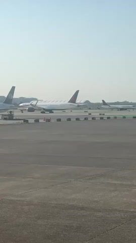 The video shows an airport tarmac on a clear day. Several large passenger airplanes are parked in the distance, including one with a Delta Airlines logo on its tail. In the foreground, a smaller airplane is taxiing on a runway. The tarmac is paved with asphalt, and there are lines and markers on the runway. The sky is blue and clear, with a few clouds visible.