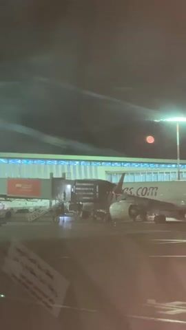 The video captures a nighttime scene at an airport tarmac in Nouaceur, Morocco. A white airplane, identified as Pegasus Airlines by the visible "s.com" on its fuselage, is parked at a gate, connected to a jet bridge. Ground crew personnel are active around the jet bridge and underneath the aircraft, suggesting ongoing boarding, disembarking, or cargo handling operations.

The airport terminal building stands in the background, illuminated by bright blue strip lighting along its roofline. Intense artificial floodlights light up the tarmac, creating a glare and a prominent red circular reflection or lens flare in the upper right portion of the frame. A partially obscured sign with visible Arabic text can be seen in the immediate foreground. The overall atmosphere is that of a bustling airport during late hours. An Arabic voiceover, likely an airport or in-flight announcement, can be heard in the audio.