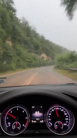 The video captures a first-person view from inside a car driving on a wet, winding road. The dashboard of the car is visible, showing the speedometer indicating speeds ranging from 86 km/h to 94 km/h. The time displayed on the dashboard is 3:27. The scene outside the car is a lush, green hillside with trees and vegetation, suggesting a rural or mountainous area. It appears to be a cloudy or overcast day, and the windshield wipers are active, indicating rain or mist. The road itself is paved and has yellow lane markings. There are orange traffic cones visible along the side of the road at one point. The overall impression is of a scenic drive through a natural landscape in potentially challenging weather conditions. The location context provided is Zaia, Bulgaria.