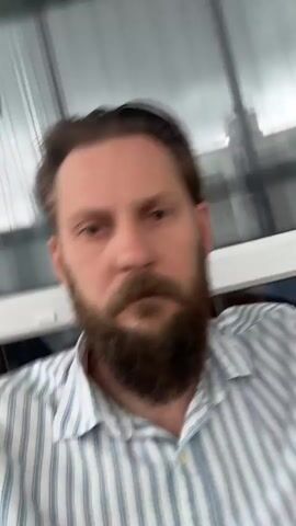 A man with a beard and brown hair is in the frame. He is wearing a blue and white striped collared shirt. The video is blurry and shaky, making it difficult to discern much of the background. The man's face is visible, and he appears to be looking at the camera. His head is moving, causing the blur in the video. There are some indistinct objects in the background, possibly a window and furniture.