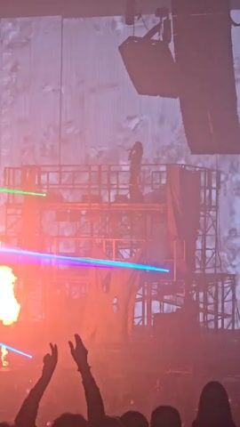 The video captures a live concert from a low angle, emphasizing the stage setup and performance. A figure is seen on a multi-tiered metal platform under bright green stage lighting. Additional beams of blue light move across the stage. The stage lights create a hazy atmosphere, and there’s a massive speaker setup hanging above the platform. The audience is visible at the bottom of the frame with raised hands, suggesting active engagement with the show. Pyrotechnics appear with flashes of orange fire on the stage, momentarily illuminating the scene.