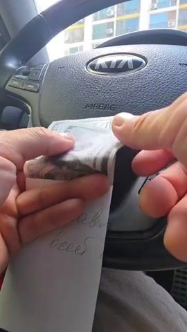 A close-up shot from inside a vehicle shows a pair of hands fanning out a wad of US hundred-dollar bills. The steering wheel of a Kia is visible in the background. A piece of paper with handwritten Cyrillic text is held beneath the money. The person is counting the money.