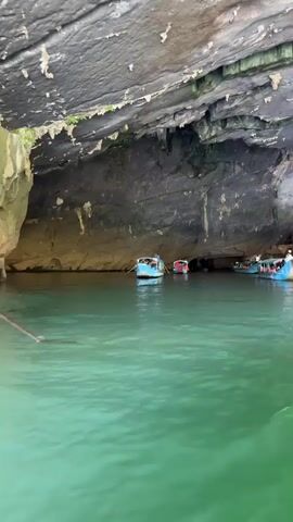 The video shows several small boats in a large cave. The cave has a rocky ceiling with stalactites and the walls are a mix of dark and tan rock. The water is a vibrant green. Several boats are visible, each with people on board. Some of the people are rowing the boats. The boats are moving slowly through the water within the cave. The scene appears to be a tourist excursion.