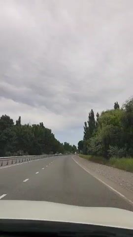 The video shows a point-of-view shot from inside a vehicle traveling on a two-lane highway. The sky is overcast with gray clouds. On the left side of the road, there is a metal guardrail and dense green trees. On the right side, there are also trees and brush, with a gravel shoulder. The road appears to be asphalt and is marked with white dashed lines. In the distance, several other vehicles can be seen on the highway. The video captures the motion of driving on the highway, with the trees and sky moving by.