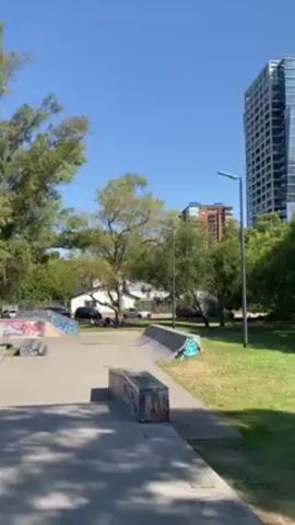 The video shows an outdoor skate park with various ramps and obstacles. There are trees and buildings in the background under a clear, bright blue sky. A young woman dressed in a white top and gray shorts enters the frame from the right and walks towards the center of the skate park.