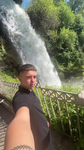 A young man stands in front of a tall waterfall, holding a camera, seemingly taking a selfie. He makes a face with his tongue sticking out in the first part of the video. He is wearing a black polo shirt and appears to be on a platform, indicated by a silver railing and lush green foliage. The waterfall roars in the background, visible from its top to its base, where it foams into a pool obscured by greenery. The weather appears sunny, and the vegetation around the waterfall is dense and green. As the camera angle shifts, the man points to the waterfall's base. Other people are walking behind the railing. The waterfall appears to be the dominant feature of the landscape.