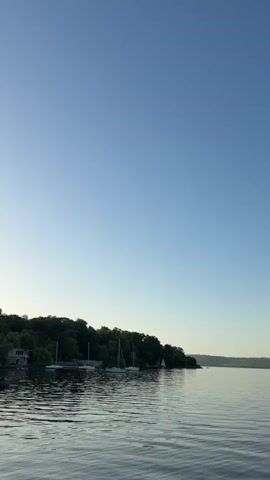 This video captures a serene waterscape with sailboats docked along a tree-lined shore. The scene is bathed in the soft light of early morning or late evening, indicated by the clear, pale blue sky that dominates the frame. Gentle ripples on the water's surface suggest calm weather. Several sailboats, with their masts visible, are moored near the shore, hinting at a leisurely day on the water. A small building is nestled amongst the trees on the left side of the frame, adding a touch of human presence to the natural setting. The distant horizon is softly defined by rolling hills under the expansive sky. The overall mood is peaceful and tranquil, inviting a sense of quiet contemplation. The location is identified as Vatra, Moldova. No visible text is present in the video.