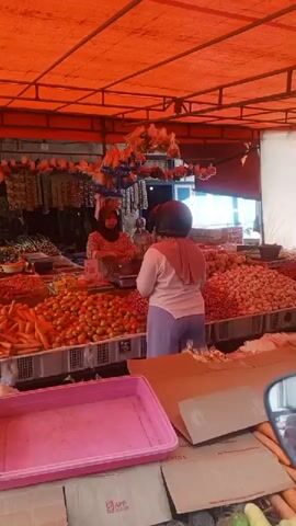 A woman wearing a pink hijab and a light-colored shirt with a helmet on is standing at a market stall. She is facing away from the camera and appears to be interacting with a vendor who is out of frame. The stall is covered by an orange tarp and is filled with produce, including tomatoes, carrots, and onions. Several other vendors and customers are visible in the background. The scene appears to be a typical outdoor market.