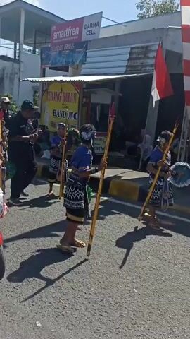 The video shows a procession or parade taking place on a paved road. In the foreground, a woman wearing a light brown hijab and a patterned top rides a red scooter. She is looking to her right. Behind her, a group of people are walking and carrying flags and what appear to be traditional instruments. One person, a young boy dressed in a blue top and a patterned skirt, is walking and holding a yellow pole. A man wearing a black shirt and a black baseball cap rides a black motorcycle in the mid-ground. He is looking to his left. Further in the background, several other people are visible, some on motorcycles and some walking. The environment appears to be a town or village, with buildings and shops lining the street. There are signs visible, including one that says "TERANG BULAN BANDUNG." The overall atmosphere seems festive and celebratory. The footage is slightly shaky, suggesting it was captured handheld.