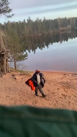 A person, possibly a child or a small adult, walks along a sandy beach towards a calm lake at what appears to be either sunrise or sunset. The sky displays soft, warm hues, which are reflected beautifully on the still surface of the water. The individual is dressed in a dark jacket and pants, a hat, and is carrying an orange life vest and another dark garment or bag. In the background, a dense forest of coniferous trees lines the far bank of the lake, their reflections visible in the water. The immediate foreground shows a portion of a green object, possibly a tent or sleeping bag, from which the video is filmed. The scene suggests a peaceful natural setting, likely in Vyritsa, Russia, with the person heading towards the water for an activity. No text is visible.