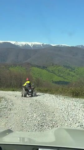 The video, shot from a moving vehicle, shows a person riding a four-wheeled ATV on a gravel road that cuts through a forest landscape. The ATV rider wears a yellow jacket and a red helmet. The vehicle recording the scene is following the ATV. The road stretches away from the camera, ascending slightly into the distance. The surrounding environment includes a dense forest, with trees ranging from bare to fully leaved. In the far background, prominent mountain peaks are visible, capped with snow. The sky is clear and blue.