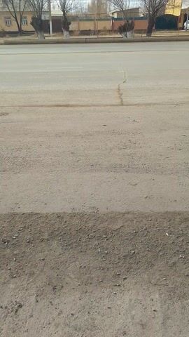 The video shows a side view of a street with a sidewalk and buildings on the left side. The road is light gray with a few cracks. To the right of the road is a darker, gravelly area.  Trees are planted along the sidewalk.  The video is shot from a high angle. It is stationary.