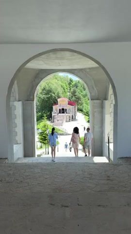 The video shows a view from under a large archway looking down a set of stone stairs. At the bottom of the stairs, three people are walking away from the camera. A woman in a light blue shirt and shorts is in front, followed by a woman in a light pink dress and a man in a gray t-shirt and shorts. Beyond them, a white church with a red roof sits on a hill surrounded by green trees. There are other people walking around the church and a white van parked in the distance. The sky is blue with some white clouds. The video pans slightly to the right, revealing more of the archway and the surrounding buildings.
