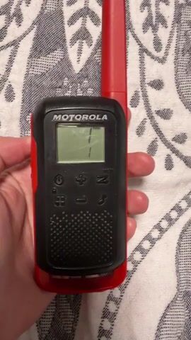 A red and black Motorola walkie-talkie is held in a person's hand against a patterned cloth background. The walkie-talkie has a small screen displaying the number 1 and "RX". The person's thumb is visible on the right side of the device. The walkie-talkie is oriented vertically with the antenna at the top.
