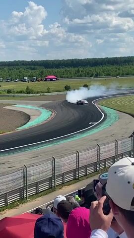 A drift car is shown on a race track, creating a large cloud of smoke as it slides around a corner. Spectators are visible in the foreground, some holding cameras and phones to capture the event. The track is surrounded by green grass and trees, with a blue sky and scattered clouds overhead. Several tents and vehicles are parked in the background.