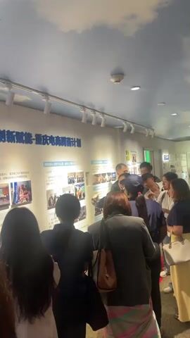A group of people are gathered around a wall display in what appears to be an exhibition or museum. The wall features text and images, suggesting a presentation or informational exhibit. One woman, facing the camera, is actively gesturing and speaking, likely explaining the content of the display to the onlookers. The crowd consists of individuals of various ages and genders, with most facing the wall and paying attention to the speaker. The environment has a clean, modern aesthetic with a light blue ceiling and track lighting. A smoke detector is visible on the ceiling. The overall scene depicts an educational or informative gathering.