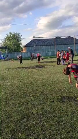 This video captures a group of children practicing soccer on a grassy field in Alkhan-Yurt, Russia. The scene appears to be an outdoor sports training session, likely for a youth soccer team.

**Subjects:** The primary subjects are a group of children, varying in age, actively engaged in soccer drills. There is also at least one adult figure, possibly a coach, observing and interacting with the children. Various soccer balls are visible on the field. The setting includes a grassy field, a fence in the background, some trees, and a building behind the fence.

**Activity/Event:** The children are participating in a soccer practice. They are seen running, kicking, and interacting with the ball. The drills appear to be focused on individual ball control and possibly some team coordination.

**Notable Details:**
*   **Time of Day:** The lighting suggests it is late afternoon or early evening, with the sun casting long shadows and illuminating the sky with soft clouds.
*   **Weather:** The sky is partly cloudy, indicating fair weather suitable for outdoor sports.
*   **Location Cues:** The presence of a soccer field, fences, trees, and a building are typical of a sports complex or schoolyard. The mention of Alkhan-Yurt, Russia provides the geographical context.
*   **Emotions:** The children appear energetic and focused on their activity, suggesting engagement and possibly enjoyment of the practice.

**Visible Text:** No discernible text is visible in the video.