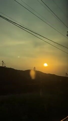 The video shows a sunset over a hilly landscape. The sun is a bright orange circle low in the sky, casting a warm glow. The sky is a gradient of yellow and blue, with a few clouds visible. In the foreground, there are silhouettes of trees and bushes. There are also power lines stretching across the frame. The video appears to have been shot from a moving vehicle, possibly a train or car, as the perspective shifts slightly throughout. There are no people visible in the video. The overall mood is peaceful and serene.