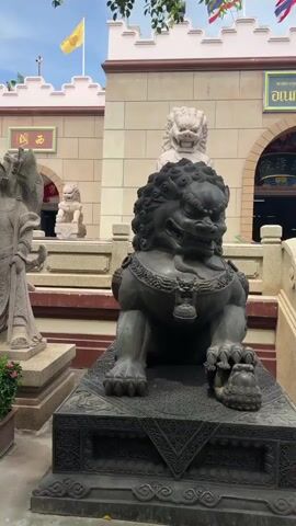 A video pans across the exterior of a temple. In the foreground, a large, dark stone lion statue sits on a pedestal. Behind it, two large stone statues of men in traditional clothing stand on separate pedestals, flanked by planters filled with pink and green foliage. Further back, a temple building with a light-colored stone facade and a pink-trimmed crenelated roof is visible. Two yellow flags fly from poles on the roof. Several smaller lion statues are placed along the steps leading up to the temple entrance. The sky is blue with some scattered clouds.