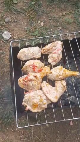 A close-up overhead shot shows chicken pieces on a grill over hot coals. The chicken is coated in a white marinade with some red pieces, possibly peppers or tomatoes. Smoke rises from the grill. The grill is set in a black metal brazier, and the surrounding ground is covered in dirt and sparse green vegetation. In the background, a shovel can be seen leaning against something out of frame. The video is shot in daylight.